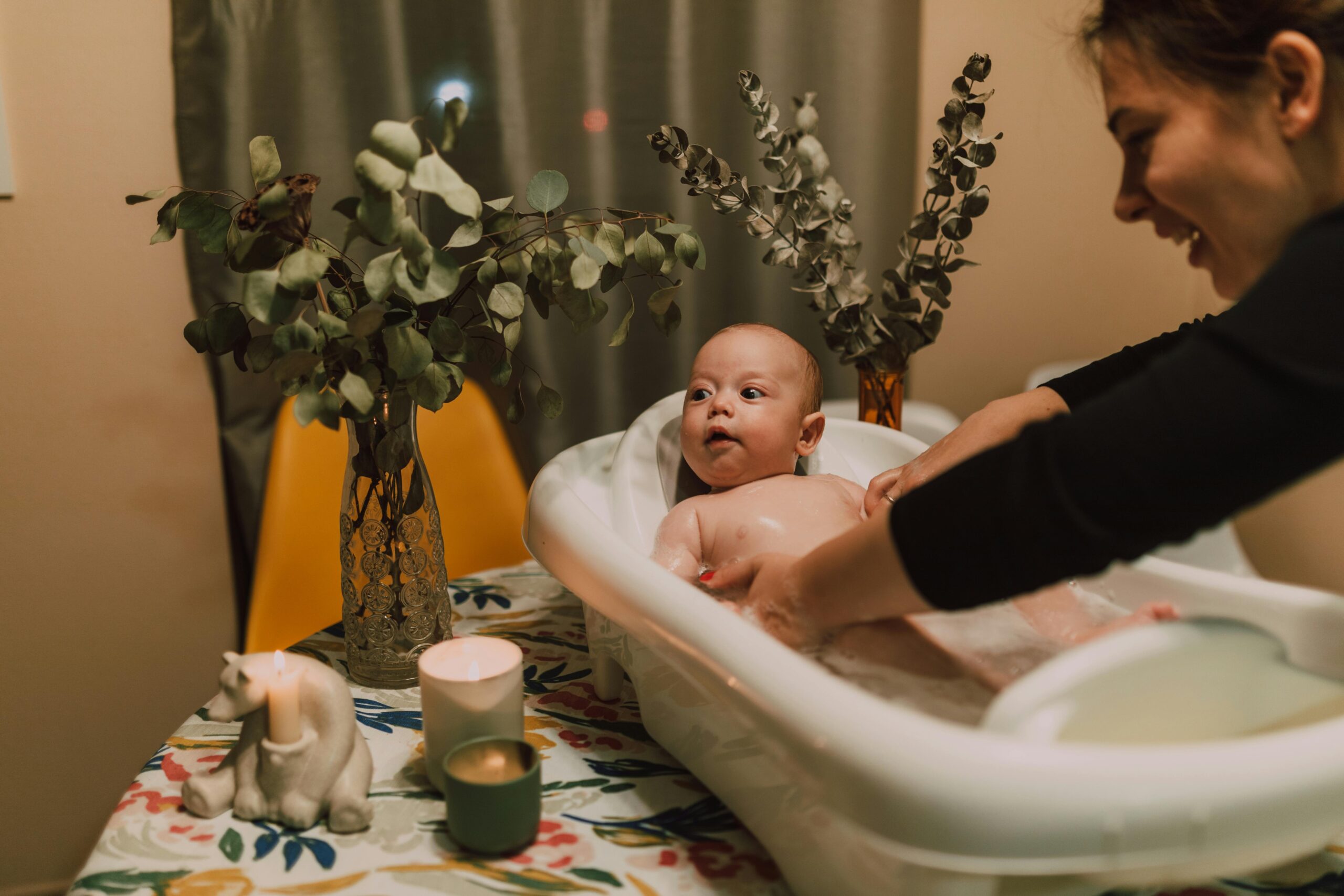 A mother lovingly baths her baby in a small tub surrounded by warm lights and plants.